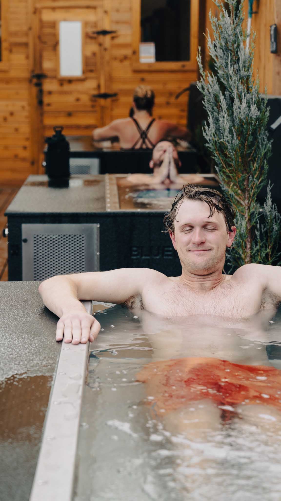 Man smiling in a BlueCube C3 cold plunge at Hooga House with two other guests plunging in the background